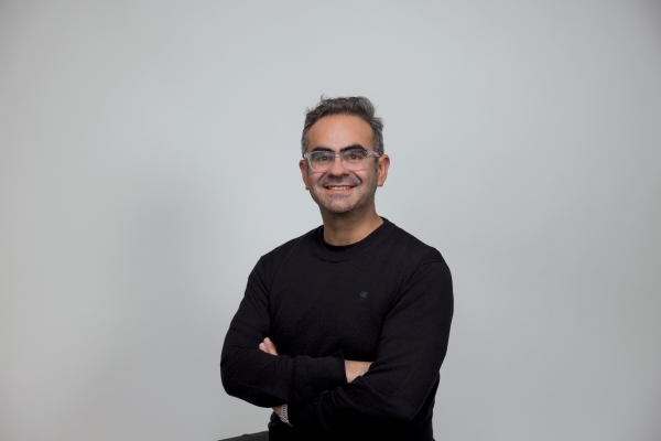 Luciano Vergara headshot, wearing glasses and a black shirt, posing with a smile and arms crossed. In front of a grey background.