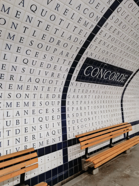 Inside a Paris metro station. The walls are covered in white tiles with a black serif letter centered in each tile. In white on a black background, the name of the station: "CONCORDE." Wooden benches line the wall.