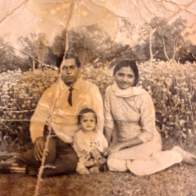 Neeraj as a child, pictured sitting on the ground in the grass at the base of a mountain with his parents sitting behind him. The photo is sepia toned.