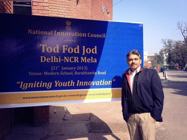 Photo of Neeraj Gulati at the Modern School in 2013 in front of a large sign advertising the presentation of Tod Fod Jod, Delhi-NCR Mela, "Igniting Youth Innovation."