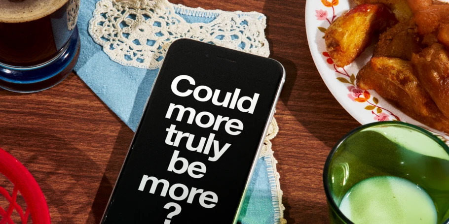 Image of phone on table with breakfast spread. Screen on phone is black with white text: "Could more truly be more?"